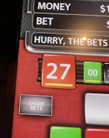 A digital display screen showing a casino betting interface, with text prompting to hurry bets and a highlighted number 27 in red. There are options for betting amounts and a 'share bets' button.