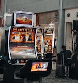 A collection of slot machines is situated inside a building with bright, colorful screens displaying various games and gaming themes. A person is seated nearby, seemingly relaxed with luggage beside them, and the atmosphere is lively with electronic displays.
